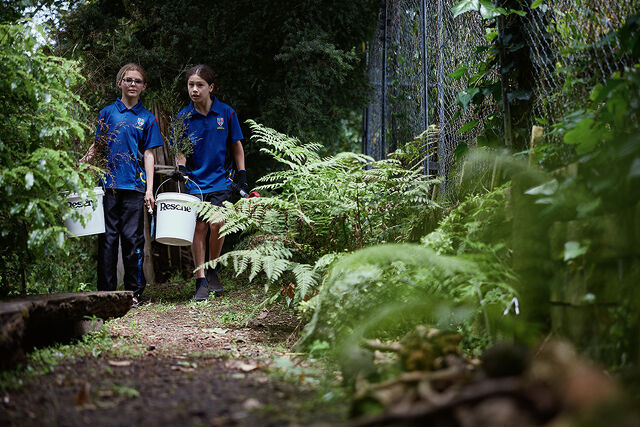 Two students carrying buckets of plants along a garden path.