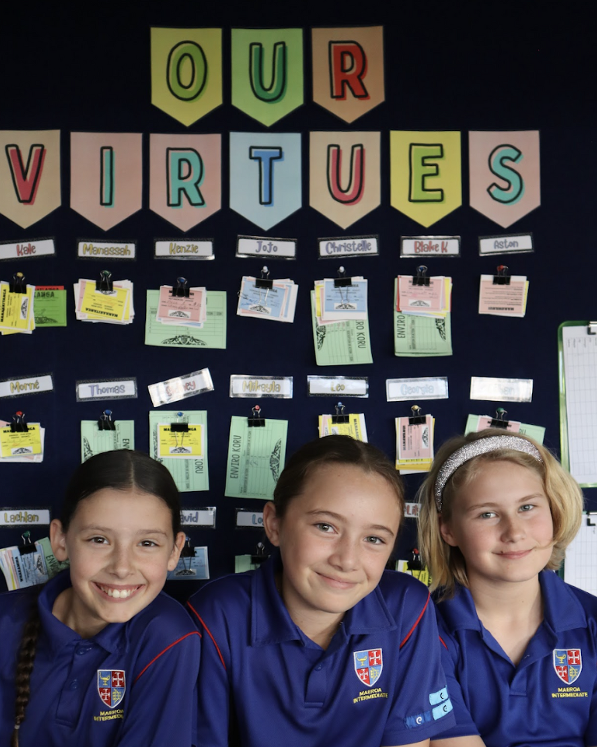 Three Maeroa Intermediate students smiling in front of a classroom display titled "Our Virtues".